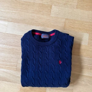 Marinblå kabelstickad tröja Morris S - Snygg marinblå kabelstickad tröja från Morris i 100% merinoull. Tröjan har rund hals, lång ärm och en röd broderad logga på bröstet.  Passar även perfekt nu inför vintern då den är ruggigt snygg och stilren. Storleken på tröjan är L men av förra ägaren har den nog krympt lite i tvätten så den sitter som S. Om du har några andra funderingar är det bara att höra av sig och priset kan diskuteras lite🫡