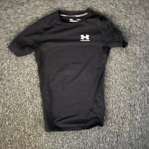 Svart compresion t-shirt från Under Armour - Svart kompressionst-shirt från Under Armour med korta ärmar och logga på bröstet. Tillverkad i stretchigt syntetmaterial som andas, perfekt för träning. Snygg och sportig passform som sitter tight mot kroppen. Lite sliten på patchen på ryggen annars bra skick.