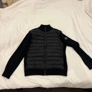 Svart pufferjacka canada goose - Svart pufferjacka från canada goose med quiltad front och stickade ärmar. Jackan har dragkedja hela vägen och en rund canada goose-logga på vänster ärm. Perfekt för dig som gillar stilren design och vill ha något som funkar till många outfits.