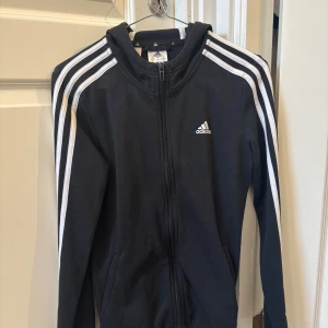 Svart Adidas hoodie med dragkedja - Svart hoodie från Adidas med klassiska vita ränder längs ärmarna och logga på bröstet. Använd ett fåtal gånger!🤗