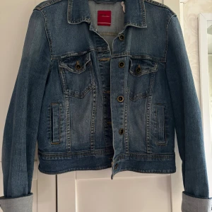 Blå jeansjacka från Divided - Snygg klassisk blå jeansjacka från Divided med knappar framtill och två bröstfickor med lock. Jackan har lång ärm och coola sömmar som ger en schysst streetkänsla. Perfekt att slänga över en hoodie eller t-shirt för en chill look.Stl 42, obetydligt använd
