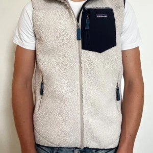Beige fleeceväst från Patagonia - Mysig beige fleeceväst från Patagonia med marinblå bröstficka och dragkedjor. Västen har hög krage och två sidofickor med dragkedja. Perfekt för lager-på-lager och chill stil.