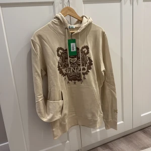 Beige hoodie med tiger från Kenzo - Snygg beige hoodie från Kenzo med ikoniskt brunt tigerbroderi på bröstet och Kenzo Paris-text. Tröjan har huva med snörning, känguruficka och ribbade muddar. Perfekt för dig som gillar streetwear och vill sticka ut med ett coolt motiv.