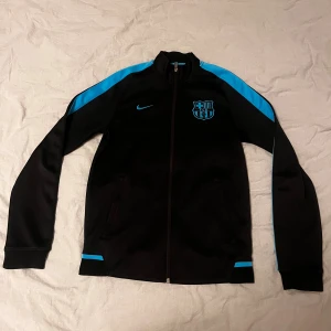 Svart Nike FC Barcelona zip hoodie - Säljer en svart FC Barcelona zip hoodie från Nike med blå detaljer på axlar och ärmar. Jackan har klubbmärket och Nike-loggan på bröstet, hel dragkedja och två fickor. Perfekt för dig som älskar fotboll och vill ha en sportig look.