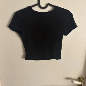 Svart croppad topp från FSBN SISTER - Svart croppad topp från FSBN SISTER i ribbad bomull. Modellen har rund hals och korta ärmar, perfekt för dig som gillar en enkel men snygg look. T-shirten är figurnära och passar till många olika stilar. Storlek S.