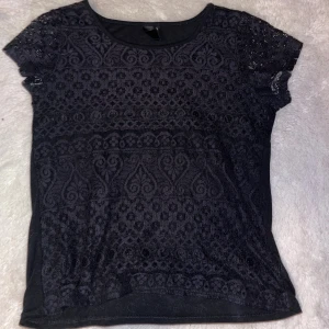 Svart spetsblus  - Svart blus från Geena i storlek M med korta ärmar och rund hals. Blusen har ett detaljerat spetsmönster över framsidan av plagget som ger en cool och elegant vibe. Materialet är mjukt och lätt, perfekt för dig som gillar att sticka ut med snygga detaljer.