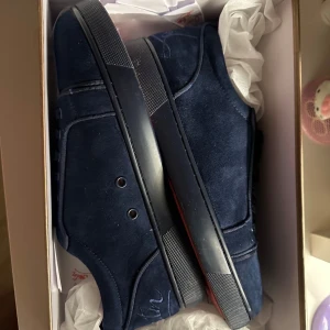 Mörkblå Louboutin sneakers i mocka - Snygga mörkblå sneakers från Christian Louboutin i mjuk mocka med klassisk röd sula och broderad logga baktill. Skorna har rund tå, snörning och diskreta detaljer som ger en lyxig känsla. Perfekta för dig som vill sticka ut med exklusiva sneakers.