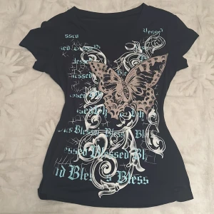 Mörkblå t-shirt med fjärilstryck - Cool mörkblå t-shirt med korta ärmar och ett stort beige fjärilstryck på framsidan. Designen har även vita och ljusblå detaljer med text och grafiska mönster. Materialet känns mjukt och stretchigt, troligtvis bomull eller polyester. Passformen är normal och figurnära.