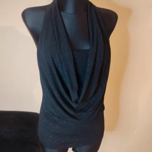 Svart glittrig halterneck topp - Svart halterneck topp med glittriga detaljer och draperad front. Toppens tyg har ett lätt skimmer och faller snyggt över bröstet. Modellen är ärmlös och har en öppen rygg, perfekt för att sticka ut med en cool look.