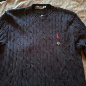 Mörkblå stickad tröja från Polo Ralph Lauren - Kabelstickad mörkblå tröja från Polo Ralph Lauren med rund halsringning och klassisk röd broderad logga på bröstet. Tröjan har långa ärmar och ribbade muddar vid ärmslut och nederkant. Perfekt för lager-på-lager under kyliga dagar.