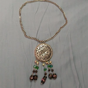 Statement halsband med stora pärlor - Unikt halsband med grov silverfärgad kedja, ca 52cm lång och stor rund medaljong med utstansat mönster,ca 6cm i diameter. Från medaljongen hänger pärlor i grönt, guld och rosa samt gröna pärlor med blommiga detaljer. Perfekt för dig som vill sticka ut med accessoarer. Pärlhängena kan tas bort