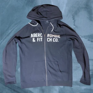 Mörkblå hoodie från Abercrombie & Fitch - Svart hoodie från Abercrombie & Fitch med vit broderad logga på bröstet. Tröjan har huva med vita snören och dragkedja framtill. Långärmad och i mjukt bomullsmaterial, perfekt för en chill och avslappnad stil. Priset kan diskuteras vid snabb affär annars är det slutgiltigt. Ha det topp//Gidlöfs