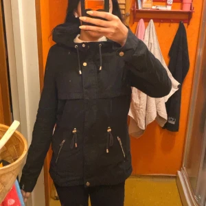 Svart parkajacka från H&M  - Svart parkajacka med huva från H&M Divided. Jackan har dragsko i midjan, dragkedja och knappar framtill samt två fickor med dragkedja och snördetaljer.
