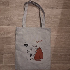Grå shoppingväska med kattmotiv - Grå shoppingväska  med tryck av två katter som tar selfie och texten 'Smile'. Väskan har långa handtag, dragkedja upptill och ett innerfack med röd dragkedja. Perfekt för att bära böcker, laptop eller annat du behöver under dagen. Mått 37x31.5cm 