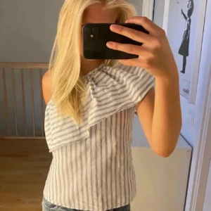 Randig offshoulder topp med volang - Säljer en fräsch offshoulder topp med ljusblå och vita ränder. Toppen har en bred volang över axeln och är ärmlös på ena sidan, vilket ger en snygg asymmetrisk look. Perfekt till jeans och sommaroutfits. Materialet känns lätt och luftigt.