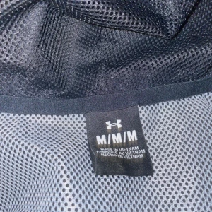Under Armour svart och vit vindjacka M - Snygg vindjacka från Under Armour i svart, vit och grått. Jackan har huva med snörning, hel dragkedja framtill och meshfoder för extra ventilation. Perfekt för dig som gillar sportig stil och vill ha en jacka som funkar när det blåser.