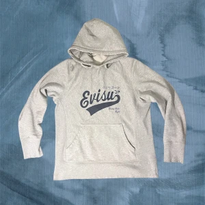 Grå hoodie från Evisu med tryck - Snygg ljusgrå hoodie från Evisu med stort mörkblått logotryck på bröstet och japanska tecken som betyder ”New York”. Mjuk bomullskvalitet som är skön att ha på sig. Perfekt för en chill och avslappnad stil. Snygg vintage slitage som ackumulerats med tiden. En av mina favoriter och vid snabb affär kan priset sänkas. Ha det gött! // Gidlöfs