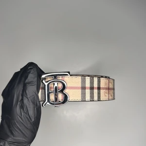 Burberry bälte svart och beach  - Snyggt Burberry-bälte i klassiskt beige rutigt mönster med svarta och röda linjer. Bältet har ett stort, silvrigt B-spänne framtill och är gjort i skinn. Perfekt accessoar för att lyfta din outfit med en lyxig touch. Om man vänder på bältet så blir det svart färg 2 färger kan man ha!