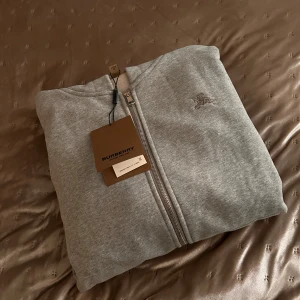 Grå hoodie från Burberry  - Grå hoodie från Burberry med dragkedja och klassisk broderad logga på bröstet. Insidan av huvan har det ikoniska rutiga Burberry-mönstret i beige, svart och rött. Tröjan är tillverkad i mjuk bomull och har huva för extra chill vibe. Står storlek XL men passar M/L