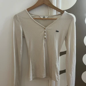 Ribbad långärmad topp från Lee - Beige ribbad långärmad topp från Lee med v-ringning och fyra små knappar framtill. Slim fit-modell med broderad Lee-logga på bröstet. Perfekt för dig som gillar en enkel men snygg stil.