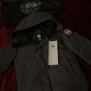 Mörkgrå dunjacka från Canada Goose - Säljer en mörkgrå dunjacka från Canada Goose med ikonisk patch på ärmen. Jackan har huva, dragkedja och knappar framtill samt två sidofickor. Perfekt för kalla dagar och har en clean, stilren look som funkar till streetwear.