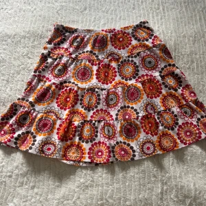 Färgglad kortkjol från me&i med volanger - Säljer en superfin kortkjol från me&i i storlek M. Kjolen har ett färgstarkt blommigt mönster i rött, orange, brunt och vitt, samt volangdetaljer som ger extra rörelse. Perfekt för dig som gillar att sticka ut med din stil!