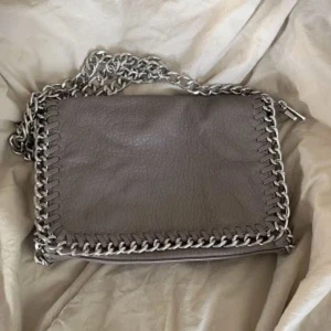 Grå/Beige Tiamo väska - Snygg grå axelväska med grov silverfärgad kedja runt kanten och som axelrem. Väskan har en rektangulär form och är tillverkad i skinnliknande material. Perfekt för dig som gillar stilrena accessoarer med lite edge, väskan är i storlek M och knappt använd. Rymlig och perfekt för vardagen 😍