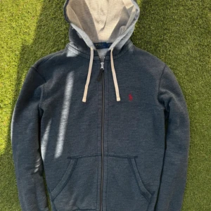 Ralph Lauren Zip Hoodie - Mörkgrå hoodie från Polo Ralph Lauren med dragkedja, vit snörning och klassisk röd logga på bröstet. Tröjan har huva med ljusgrå insida och två fickor framtill. Tillverkad i mjuk bomullsblandning, perfekt för chill dagar.