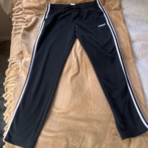 Svarta Adidas mjukisbyxor med vita ränder - Svarta mjukisbyxor från Adidas med klassiska vita ränder längs sidorna och Adidas-logga på låret. Byxorna har resår och snörning i midjan för skön passform. Perfekta för chill eller träning, tillverkade i mjukt material.