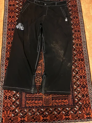 Svarta täckbyxor med vita sömmar - Svarta täckbyxor med vita kontrastsömmar och dragkedjor vid bensluten. Byxorna har fickor fram och bak samt bälteshällor. Detaljer som broderad logga och patch på ena benet. Perfekta för vinterns äventyr.