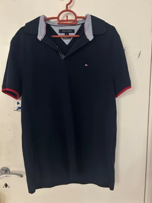 Marinblå t-shirt Tommy Hilfiger - Marinblå t-shirt från Tommy Hilfiger med klassisk krage och korta ärmar. T-shirten har röda detaljer vid ärmslut och en liten broderad logga på bröstet. Perfekt för en clean och stilren look.