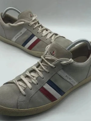 Moncler sneakers med röd och blå detalj - Snygga sneakers från Moncler i beige mocka med klassiska ränder i rött, vitt och blått på sidan. Skorna har rund tå, platt sula och snörning framtill. Moncler-logga på plösen och sidan ger en exklusiv touch. Liten fläck på ena skon, där av det billiga priset, går att få bort med skorengöringsmedel