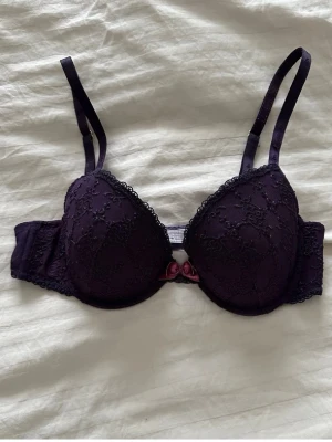 Mörklila spets-bh från Lella M - Snygg mörklila bh från Lella M med vadderade kupor och spetsdetaljer. Dekorerad med en liten rosett i vinrött framtill. Justerbara axelband och hakknäppning bak. Perfekt för dig som gillar feminina detaljer och en bekväm passform.