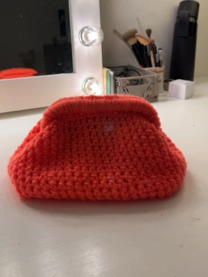 Handvirkad orange kuvertväska - Unik handvirkad kuvertväska i stark orange färg. Väskan har en mjuk, stickad struktur och en klassisk form med metallstängning upptill. Perfekt accessoar för dig som vill sticka ut med något personligt och färgglatt.💗Väskan är 9 gånger 15 skulle jag säga i metallställningen.