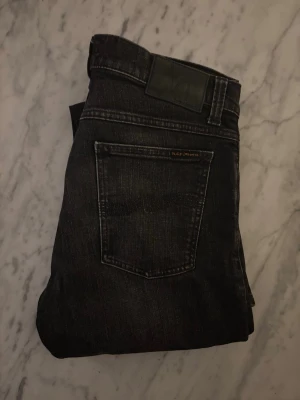 Mörk gråa jeans från Nudie Jeans - Snygga mörkgråa jeans från Nudie Jeans med klassisk femficksdesign och diskreta slitningar framtill. Jeansen har rak passform och är tillverkade i denim av bomull. Perfekta för en stilren och avslappnad look.