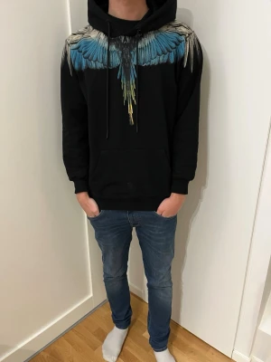 Svart Marcelo Burlon Hoodie! - Snygg svart hoodie från Marcelo Burlon med ikoniskt vingtryck i blått, vitt och gult över axlar och bröst. Modellen på bilden är ca 185!