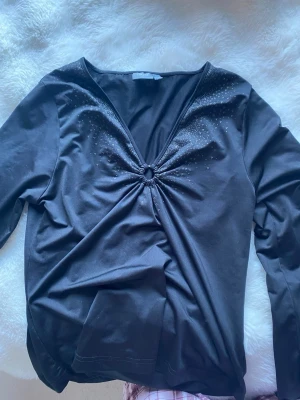 Svart v-ringad topp med ringdetalj - Snygg svart långärmad topp med v-ringning och en dekorativ ringdetalj framtill. Små glittriga stenar pryder ringningen för extra stil. Perfekt till jeans eller kjol för en trendig look.