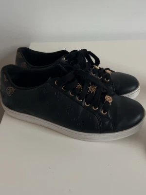 Svarta sneakers från Guess med gulddetaljer -  Svarta  Guess skor  med guldfärgade detaljer o  logomönster på sidorna. Använda bara ett par gånger . Har inga skador utan bara lite märken på undersidan (går säkert bort med tvätt) , men det är inget som syns. Köpa för 1100kr.