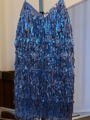 Silver  paljettklänning med fransar - Supersnygg blå kortklänning med tunna axelband och massor av glittriga paljetter och fransar som ger ett riktigt statement. Perfekt för dig som vill sticka ut och glänsa på dansgolvet. Klänningen har en v-ringning och är ärmlös.