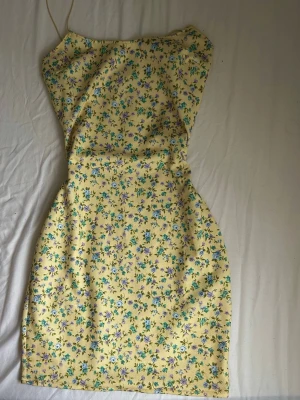 Gul blommig kortklänning - Söt gul kortklänning med smala axelband och blommigt mönster i blått, grönt och lila. Klänningen har en figurnära passform och är perfekt för varma dagar. Tillverkad i ett lätt och bekvämt material.