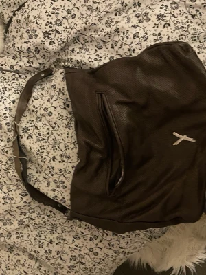 Mörkbrun handväska i skinn - Snygg mörkbrun handväska i skinn med axelrem och silverfärgade metalldetaljer. Väskan har en praktisk dragkedja framtill och ett diskret märke i form av ett X. Perfekt för dig som vill ha en stilren och rymlig väska till vardagen.