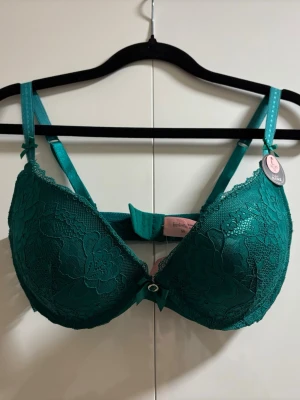 Grön spets-bh från Finesse, kupa E - Snygg grön bh från Finesse sopm har vadderade kupor och vacker spets med blommönster. Justerbara axelband och liten rosett framtill. Perfekt för dig som vill ha både stöd och stil. Kupan är E och omkretsen 70.
