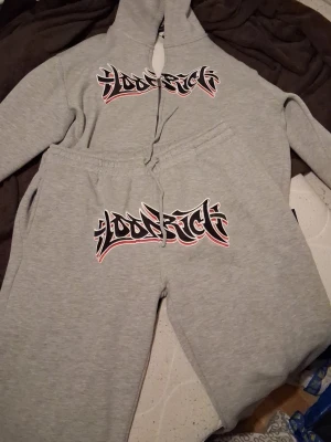 Grått Hoodrich set med tryck - Grått tvådelat set från Hoodrich med matchande hoodie och byxor. Båda delarna har ett stort svart och vitt Hoodrich-tryck med röda detaljer på bröstet och låret. Setet har sportig streetwear-stil och är gjort i mjukt material med huva och dragsko. JD exclusive (i think) kan bytas ut mot ett essentials fear of god set svart Med storlek Xl rlr L eller denim tears i samma