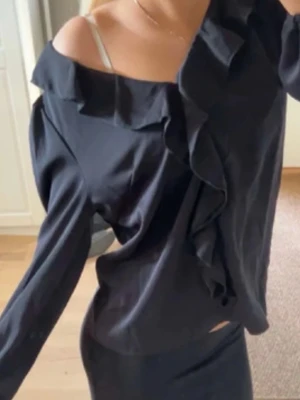 Svart blus med volangdetalj - Stilren svart blus med volang längs v-ringningen och axlarna. Blusen har långa ärmar och en lös passform som ger en avslappnad men chic look. Perfekt att matcha med kjol eller jeans för en trendig outfit.