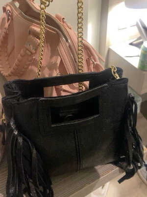 Svart handväska med fransar och kedja - Stilren svart handväska med dekorativa fransar på sidorna och guldfärgad kedjerem. Väskan har ett rektangulärt handtag upptill och är tillverkad i ett material som liknar skinn. Perfekt accessoar för att lyfta din outfit.