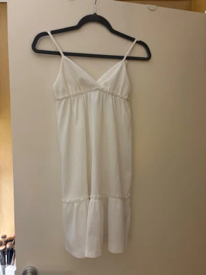 Vit sommarklänning med smala axelband - Söt vit klänning med smala axelband och v-ringning. Klänningen har en volang nertill, vilket ger en luftig känsla. Perfekt för varma dagar och enkel att styla med accessoarer. Fint skick!!!