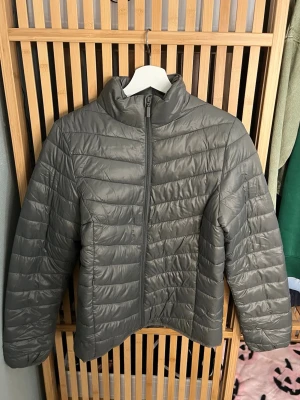 Grön pufferjacka med dragkedja - Snygg grön pufferjacka med hög krage och hel dragkedja framtill. Jackan har quiltad design och långa ärmar, perfekt för kyligare dagar. Enkel och stilren modell som passar till många outfits.