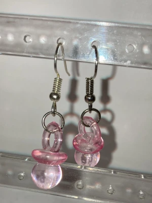Rosa nappörhängen i plast - Handgjorda unika örhängen med nappformade hängen i genomskinlig rosa plast. Krokar i metall ger en cool kontrast till de lekfulla detaljerna. Perfekta för dig som vill sticka ut med accessoarer som har en twist. Kontakta mig gärna om du har frågor eller vill köpa.