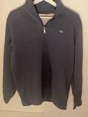 Blå halvzip sweatshirt från Gant - Mörkblå sweatshirt från Gant med halv dragkedja och liten broderad logga på bröstet. Tröjan har hög krage, långa ärmar och ribbade muddar. Perfekt för en avslappnad och stilren look.