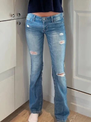 Blå bootcut jeans med slitningar - Snygga ljusblå bootcut jeans med slitna detaljer och broderade bakfickor. Jeansen har låg midja och klassisk femficksdesign. Perfekta för en avslappnad och trendig look.